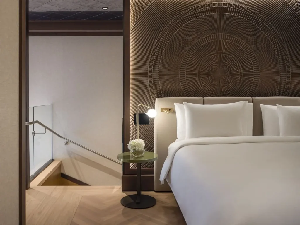 Eine moderne Duplex-Suite verfügt über ein weißes Bett, einen runden Beistelltisch mit einem Blumenarrangement, ein geometrisches Holzkopfteil und eine Treppe mit einem Glasgeländer im Hintergrund.